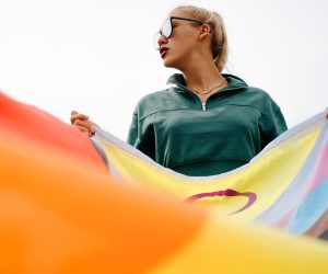 Fashionable young woman with glasses and red lipstick holds rainbow flag, international symbol of LGBTQ+ community, gay, lesbian, people of color and transperson. Equal rights and freedoms for LGBT.
